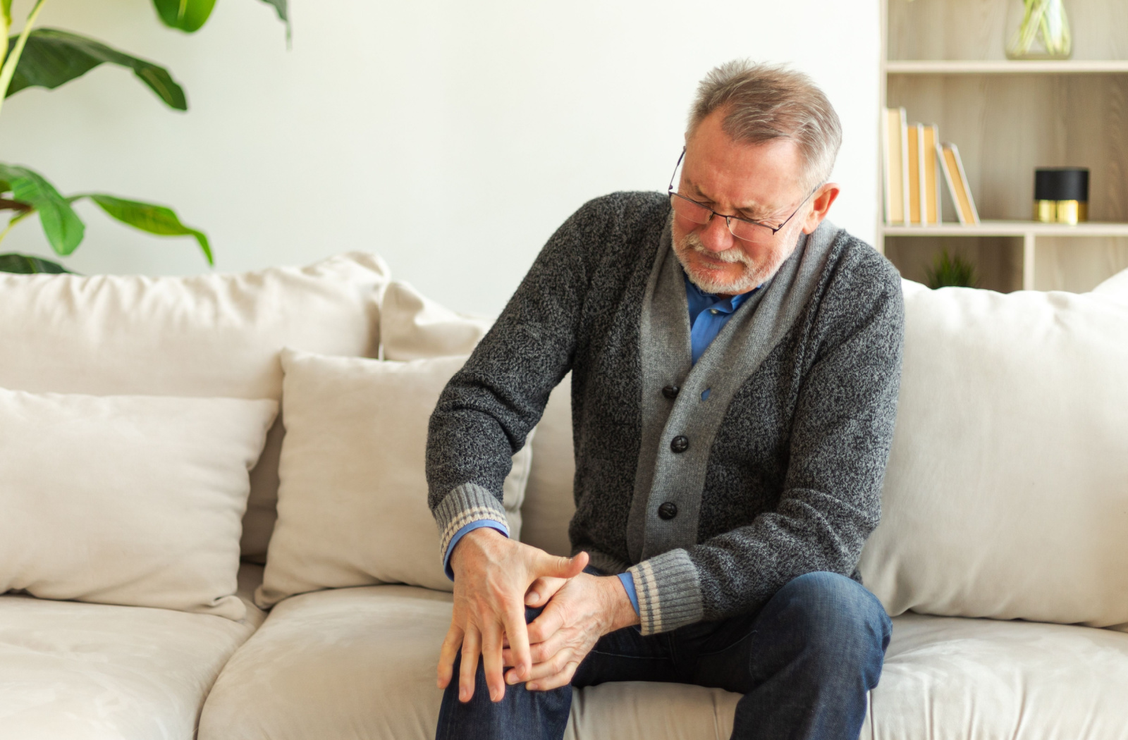 An older adult man sitting on a couch and holding his right knee due to pain

