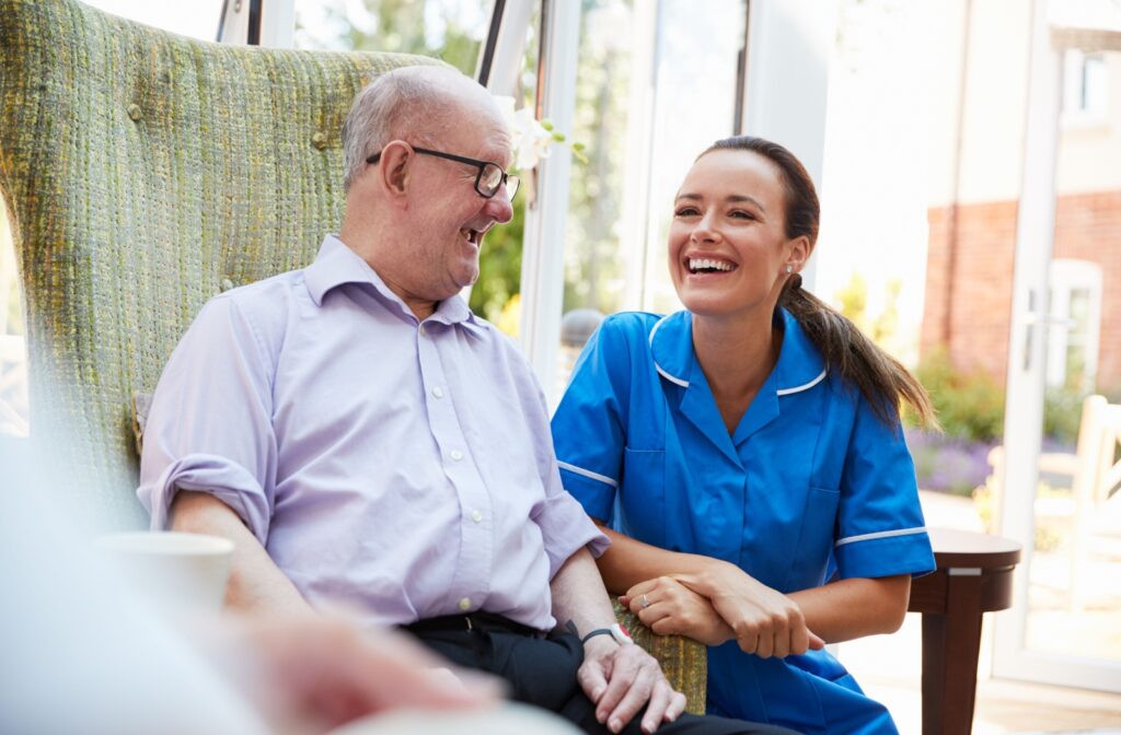 An older adult man in a memory care facility sitting on a chair smiling and having a conversation with a nurse.